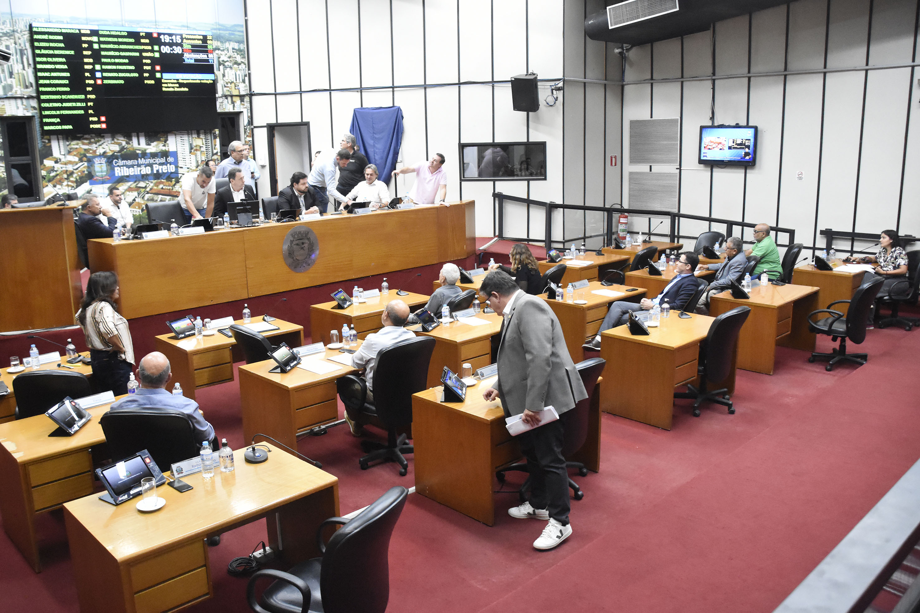 Imagem aberta do Plenário com os vereadores de costas e a Mesa Diretora de frente